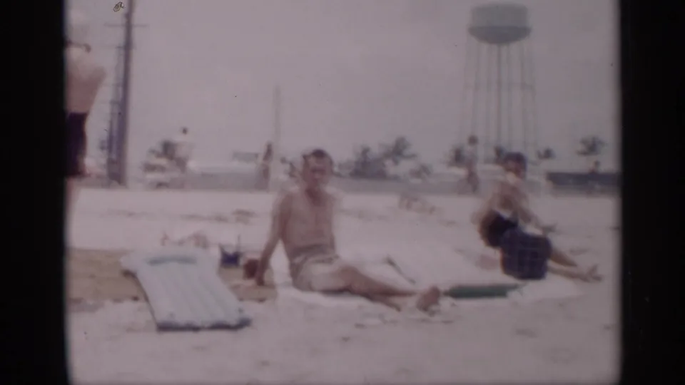 1959 California beach scene with a seated man and woman on sand, a light blue inflatable mattress, palm trees, other beachgoers, and a water tower behind them.