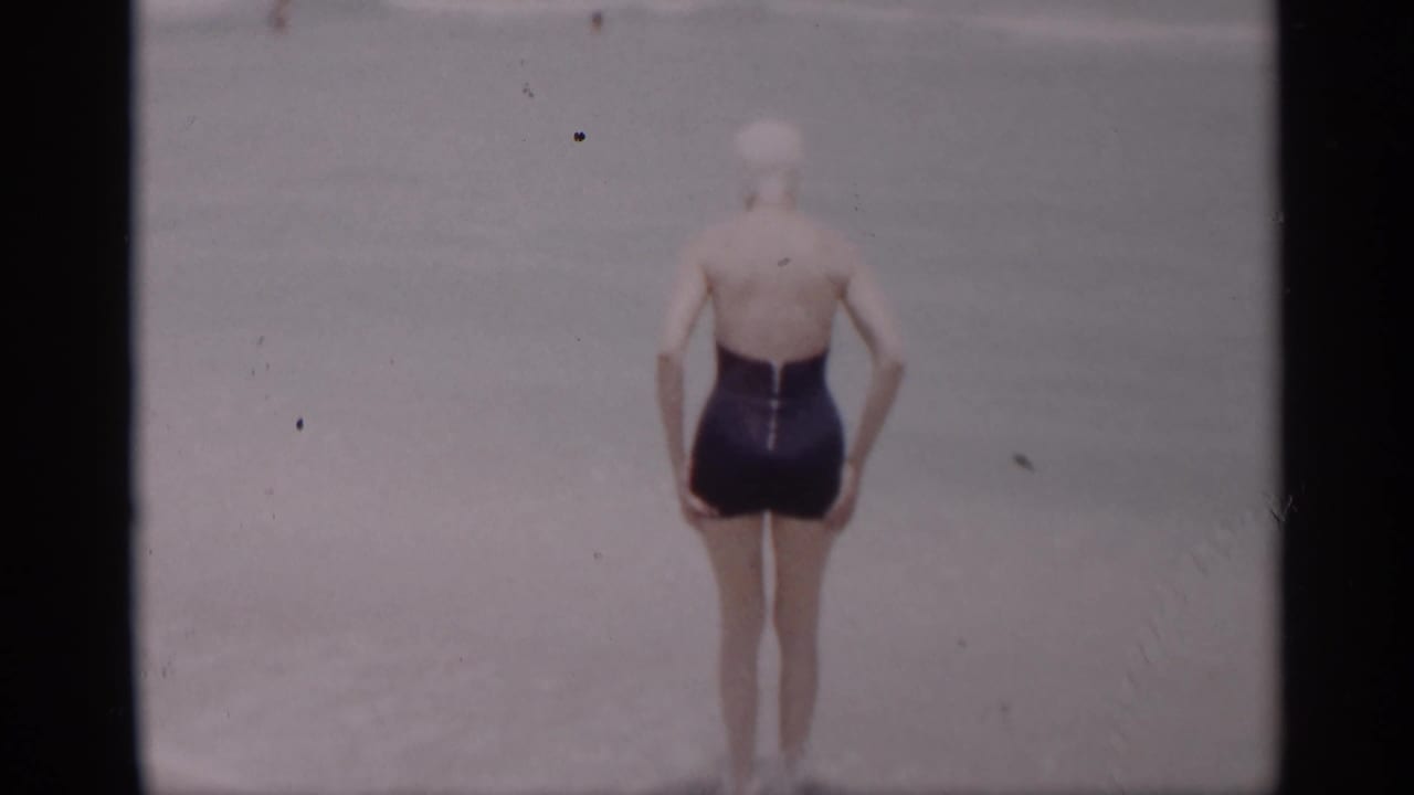 A woman in a dark one-piece swimsuit and white swim cap stands at the California ocean edge in 1959 footage.