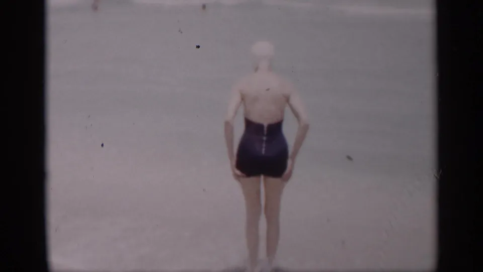 A woman in a dark one-piece swimsuit and white swim cap stands at the California ocean edge in 1959 footage.