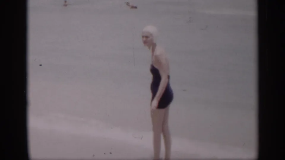 In the 1959 California beach footage, the woman turns in profile beside the pale surf and sand.