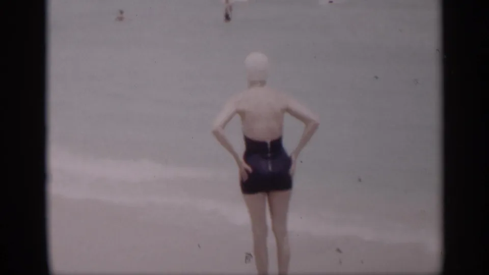 The woman in a dark swimsuit and white cap stands hands on hips, looking toward the water in 1959 California beach footage.