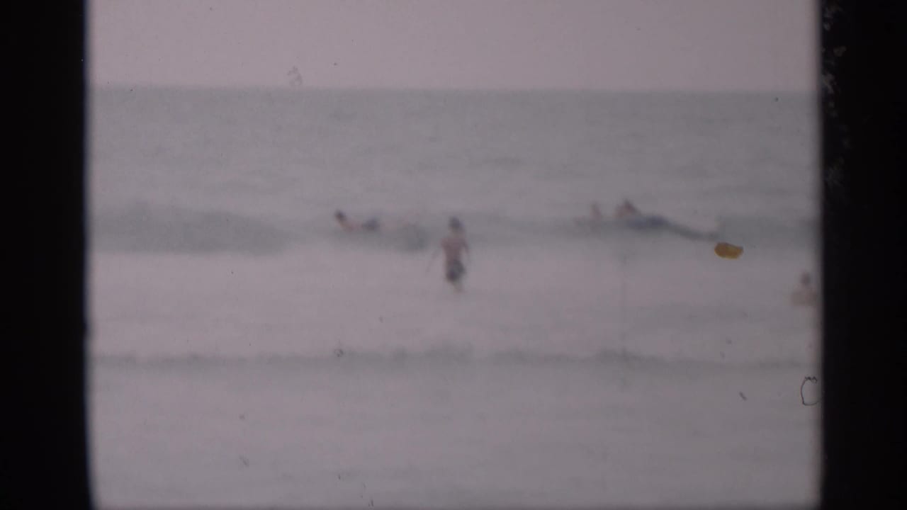 Distant swimmers stand and move in hazy ocean surf with a soft horizon behind them.