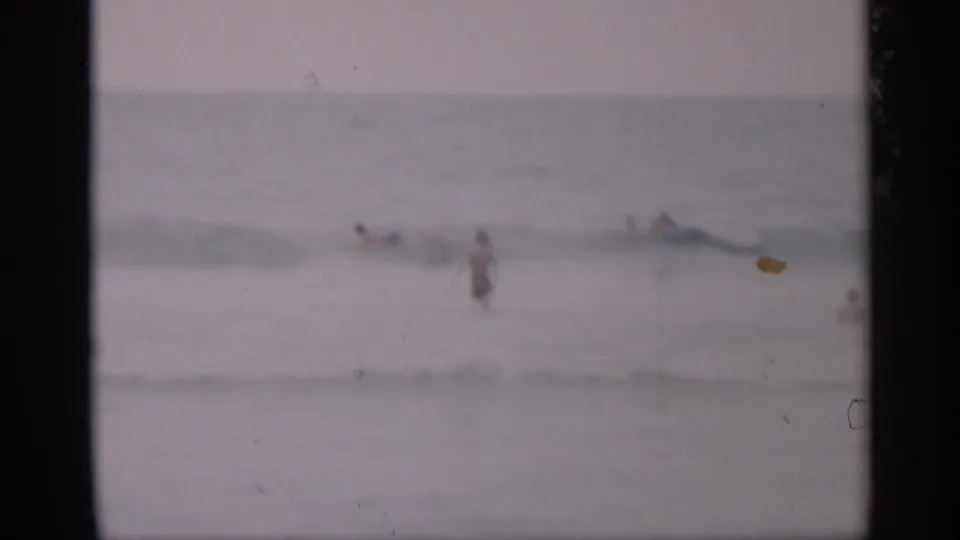 1959 California 8mm frame showing distant beachgoers wading in pale, hazy surf under an indistinct sky.