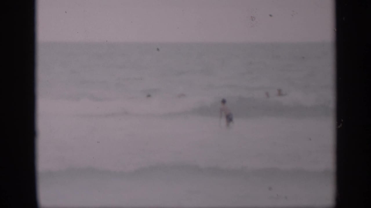 Soft 1959 California beach frame with small, distant swimmers scattered across low waves and muted ocean tones.