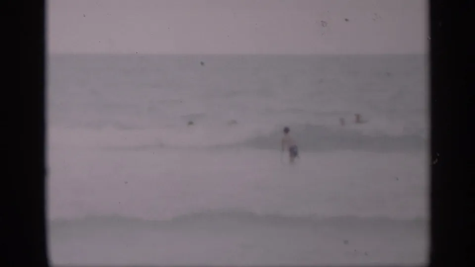 Soft 1959 California beach frame with small, distant swimmers scattered across low waves and muted ocean tones.