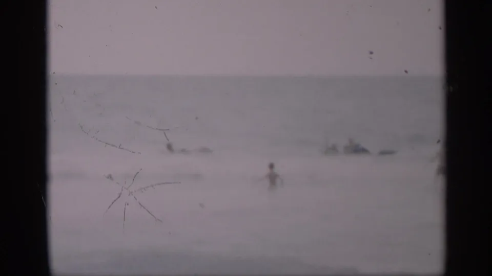Grainy 8mm view of a California beach scene with one distant wader in front of small groups in the ocean.
