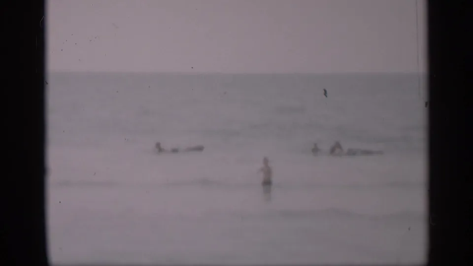1959 California 8mm frame with multiple distant beachgoers in hazy ocean water, framed by visible film borders.