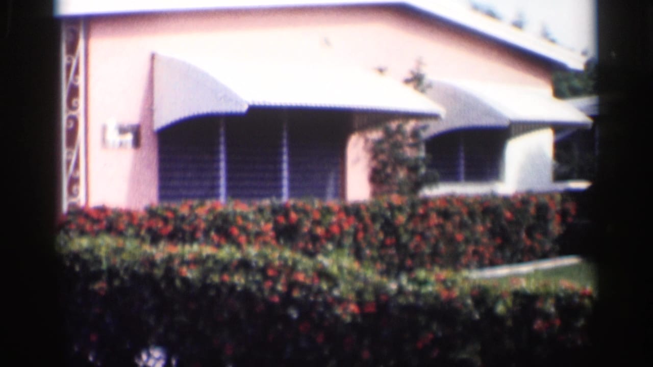 Pink house exterior with white awnings behind red rose hedges and lawn.