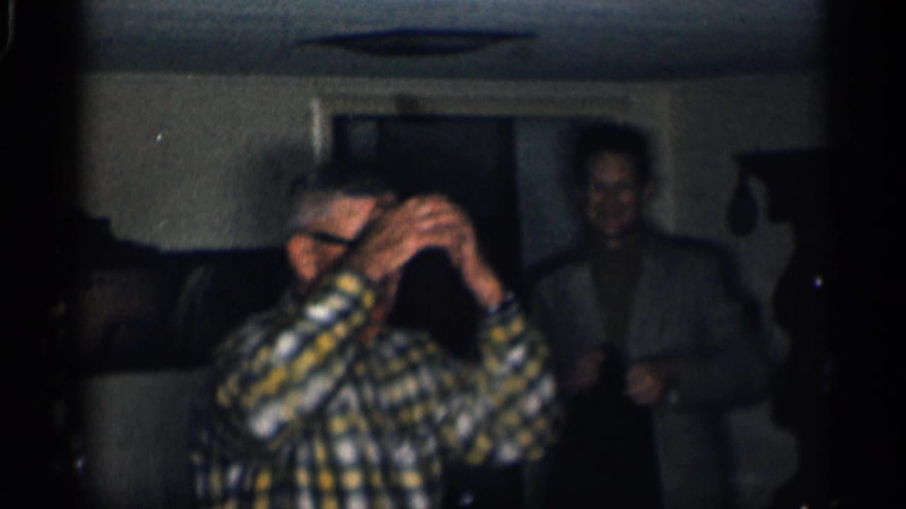 1959 California home-movie frame of adults indoors, with a man in a yellow plaid shirt lifting his hands to his glasses.