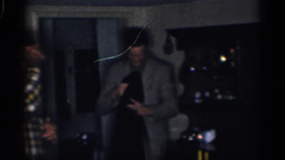 1959 California indoor gathering frame with adults standing near curtains, including a suited man holding a dark garment.