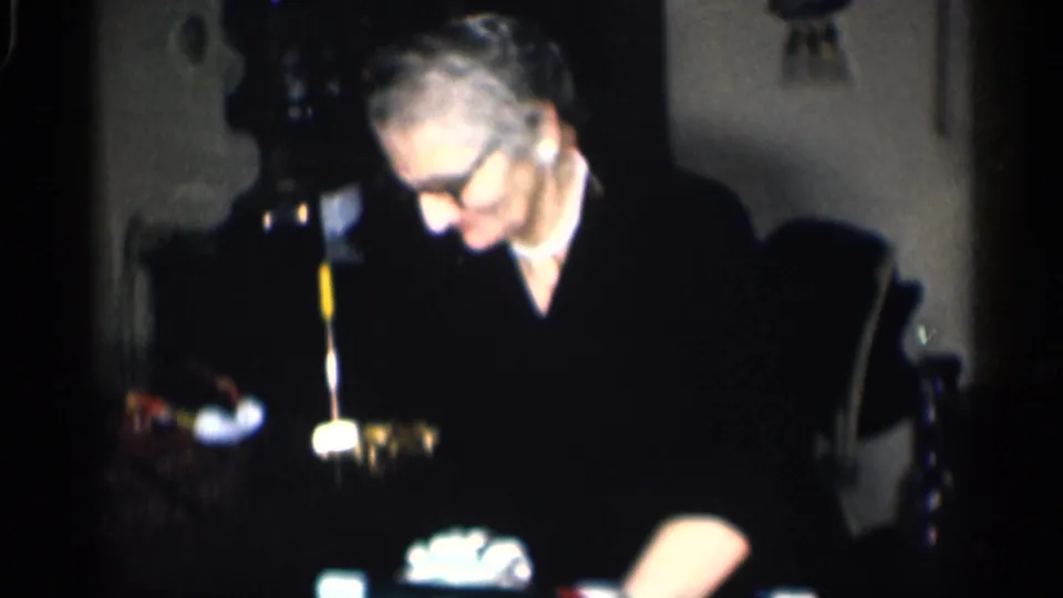 1959 California home-movie frame of an older man in glasses seated at a table inside a private home.
