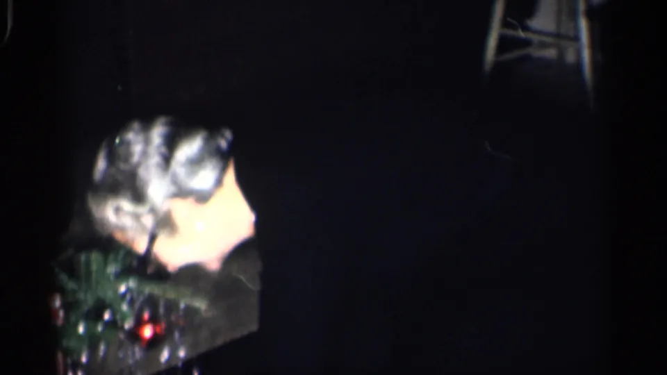 1959 California home-movie frame of an older woman with glasses seated in profile inside a dim private room.