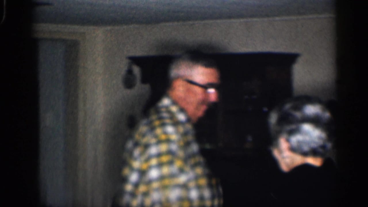Older man in plaid shirt and glasses beside an older woman indoors.