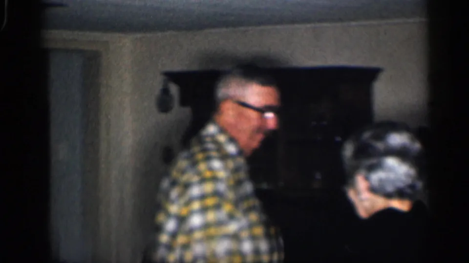 Older man in a yellow-blue plaid shirt and glasses stands near an older woman in a dim California home interior.