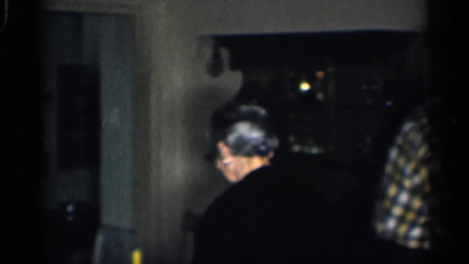 An older woman is seen from behind while an older man in plaid moves nearby in the dim home interior.