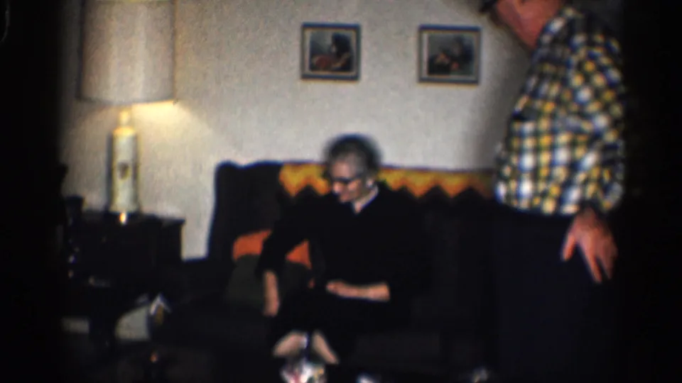 In a 1959 California living room, an older woman sits on a dark couch while an older man in plaid stands beside her.