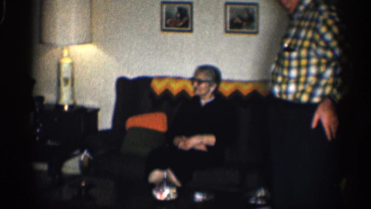Elderly woman sits on a couch while an elderly man stands nearby in a living room.