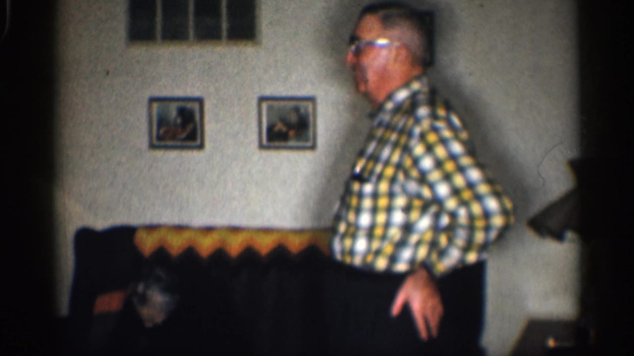 Elderly man in glasses stands near a couch in a living room with framed pictures.