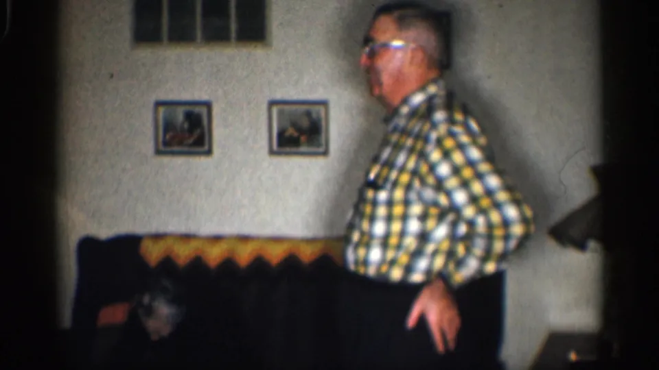1959 California home-movie frame of an elderly man in glasses and a plaid shirt standing beside a couch in a domestic interior.