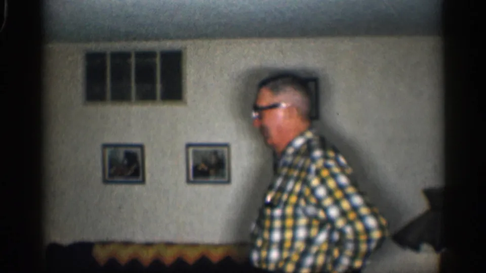 1959 California home-movie frame showing an elderly man in profile near a couch, with framed pictures on a textured wall.