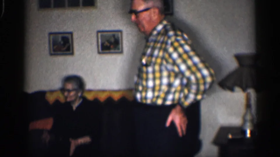 1959 California home-movie frame with an elderly man standing near an elderly woman seated on a couch in a private living room.