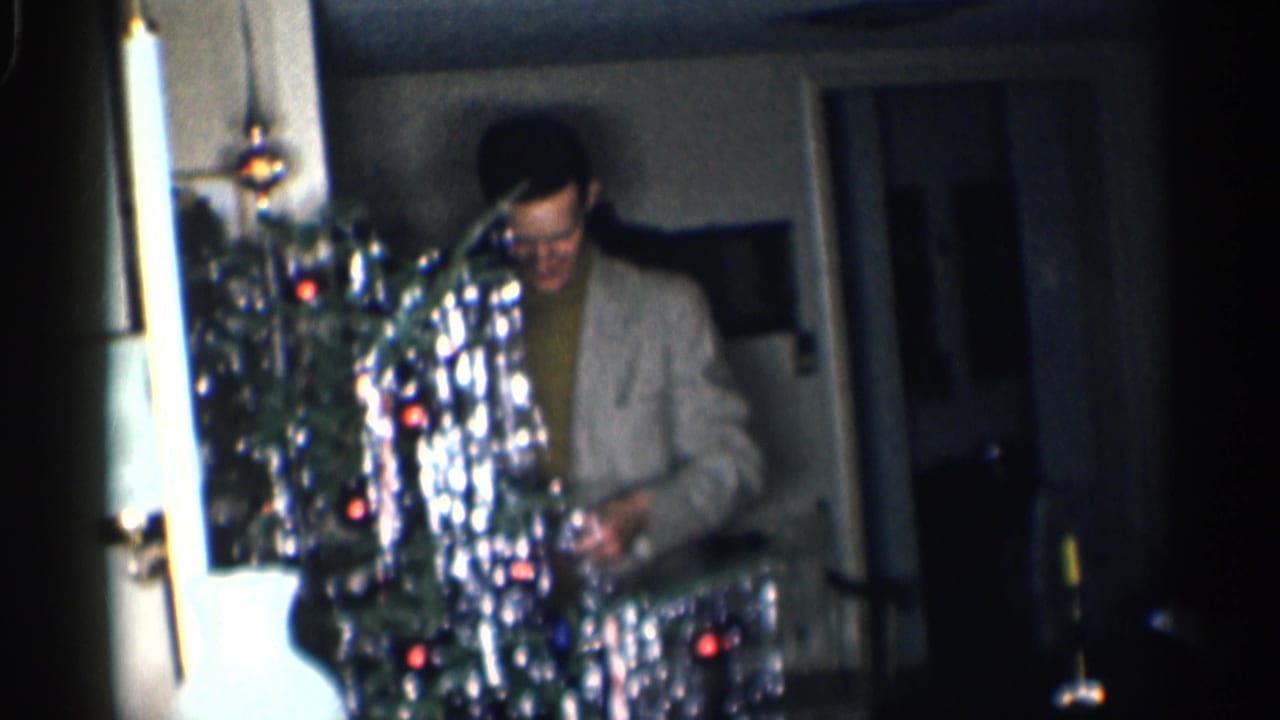 Man in glasses decorates a tinsel Christmas tree in a dim home interior.
