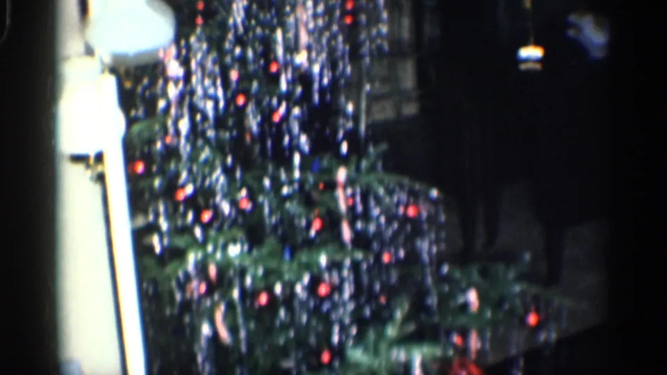 A close home-movie view shows the tinsel Christmas tree, red ornaments, and nearby lamp in a dark California interior.