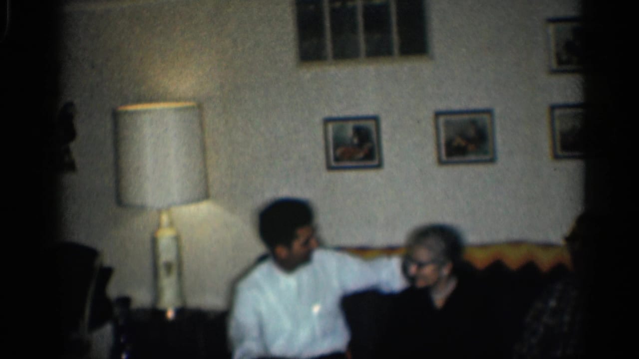 1959 California home-movie frame showing three adults seated on a patterned couch in a softly lit living room.