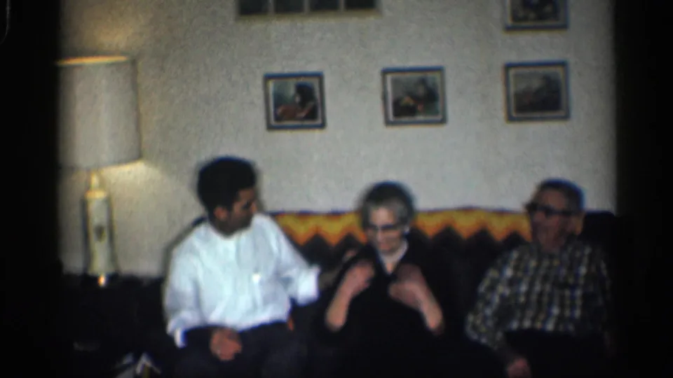 1959 California living-room frame with three adults on a brown couch with a yellow and orange patterned backrest.