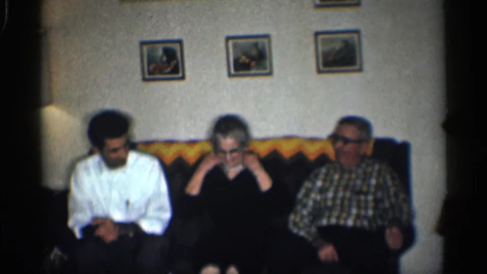 Home-movie frame from 1959 California showing a seated woman raising her hands to her hair between two men.
