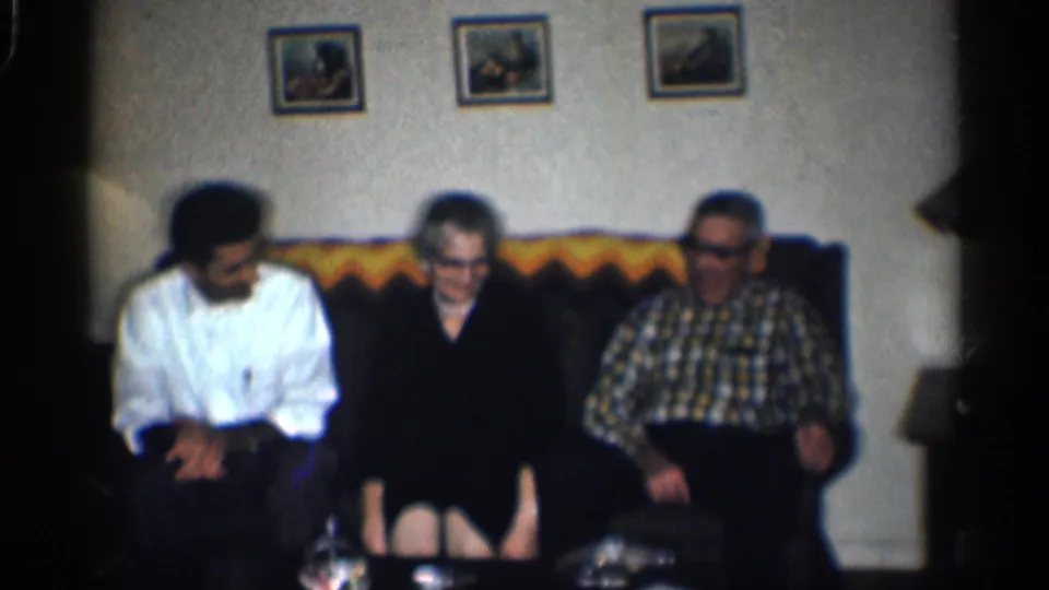 1959 California home-movie frame showing three adults seated together in a dim living room with framed wall pictures.