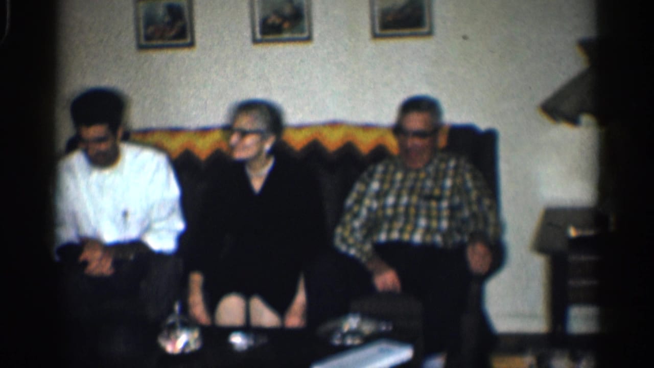 Three adults sit on a dark couch beneath framed wall pictures in a living room.