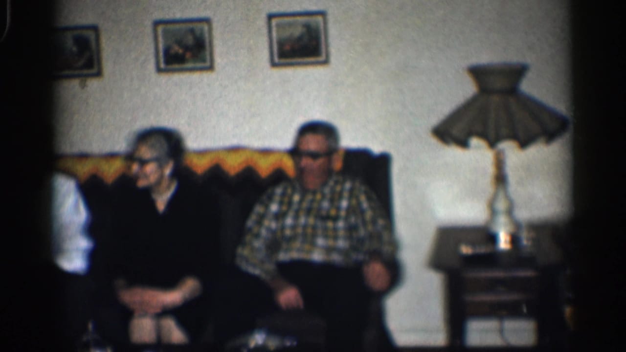 Two older adults remain seated on the couch as a man in a white shirt is partly cropped at left.