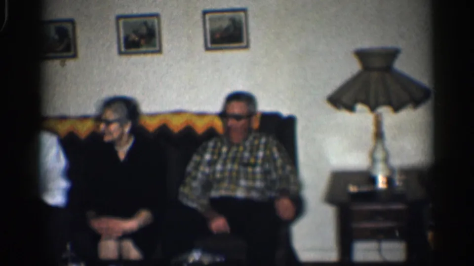1959 California living-room frame with two older adults on a couch, a partly visible younger man, framed pictures, and a side-table lamp.