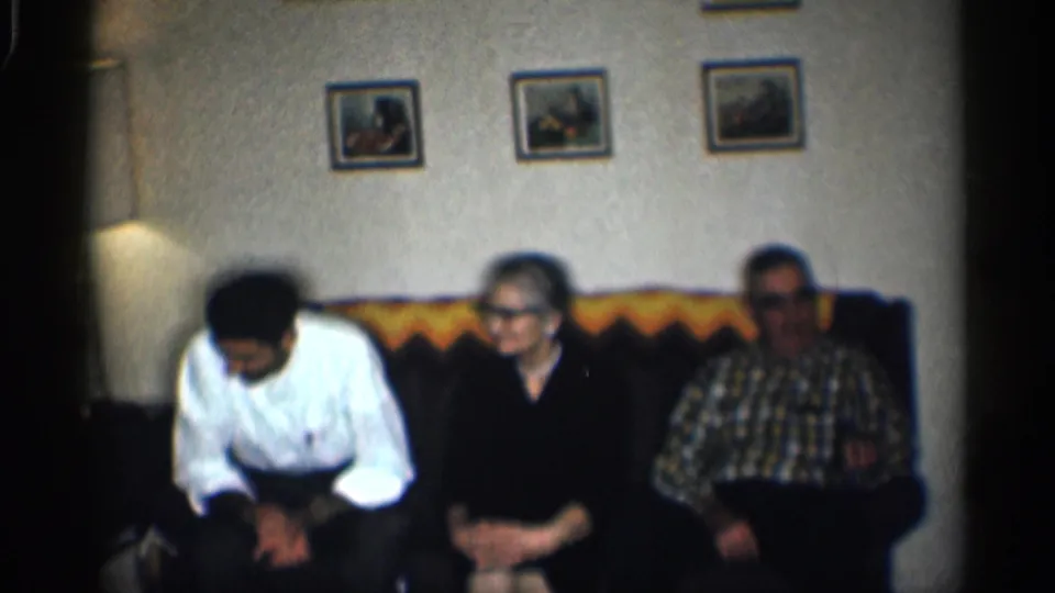 1959 California home-movie frame showing a man leaning forward beside two older adults on a couch under framed wall pictures.