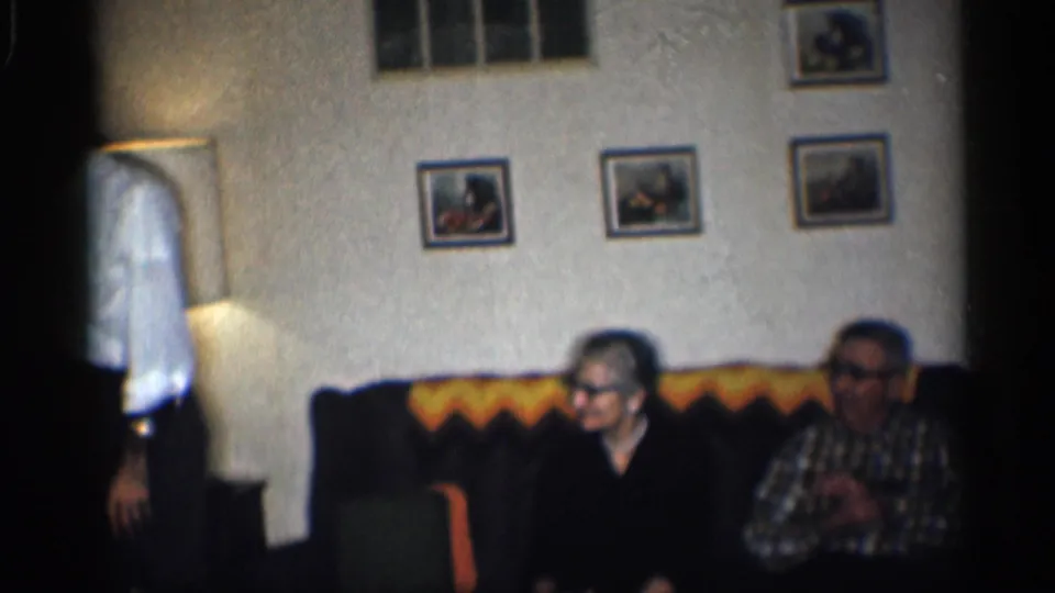 1959 California living-room frame with two older adults seated on a couch and a man partly visible standing at the left edge.