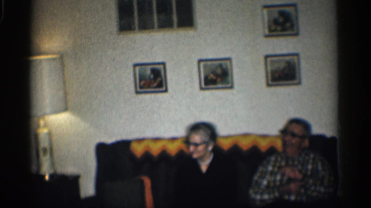 In a 1959 California home interior, an elderly woman and man sit on a dark couch under framed pictures and warm lamp light.