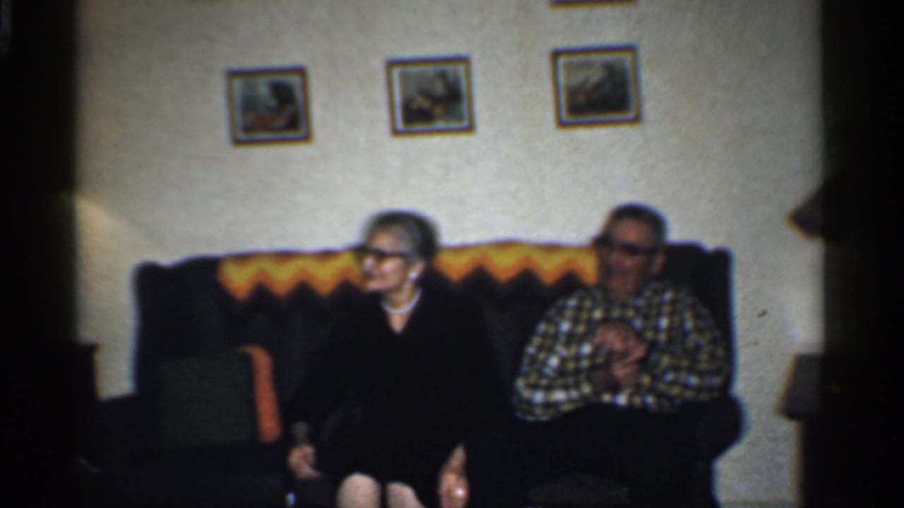 Older woman and man sitting on a couch in a living room with wall pictures.