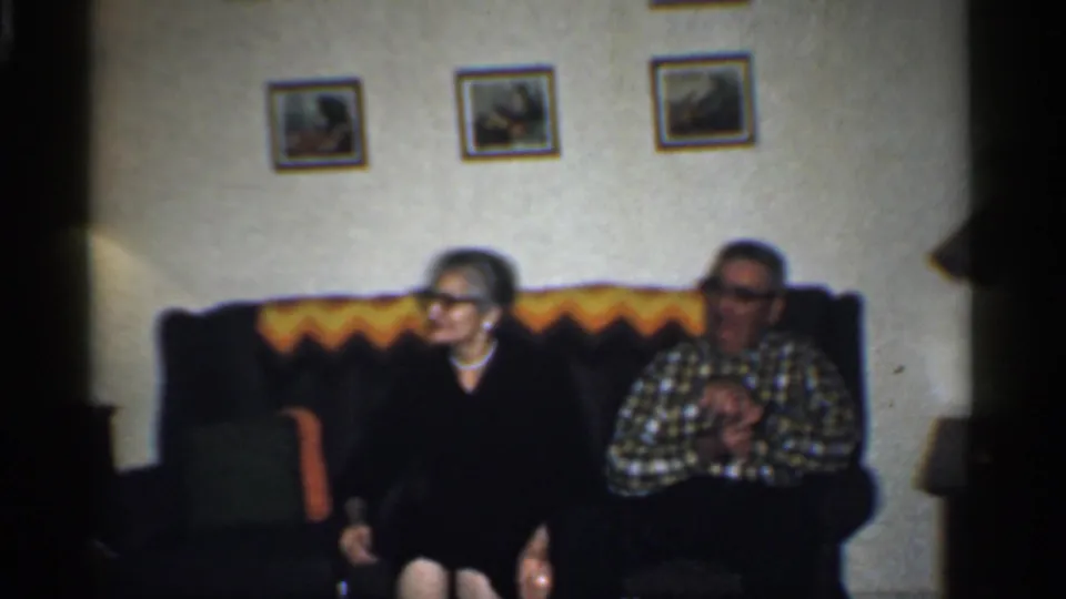 The older couple remains seated on the couch in the California living room, with framed pictures above them and a patterned throw behind them.