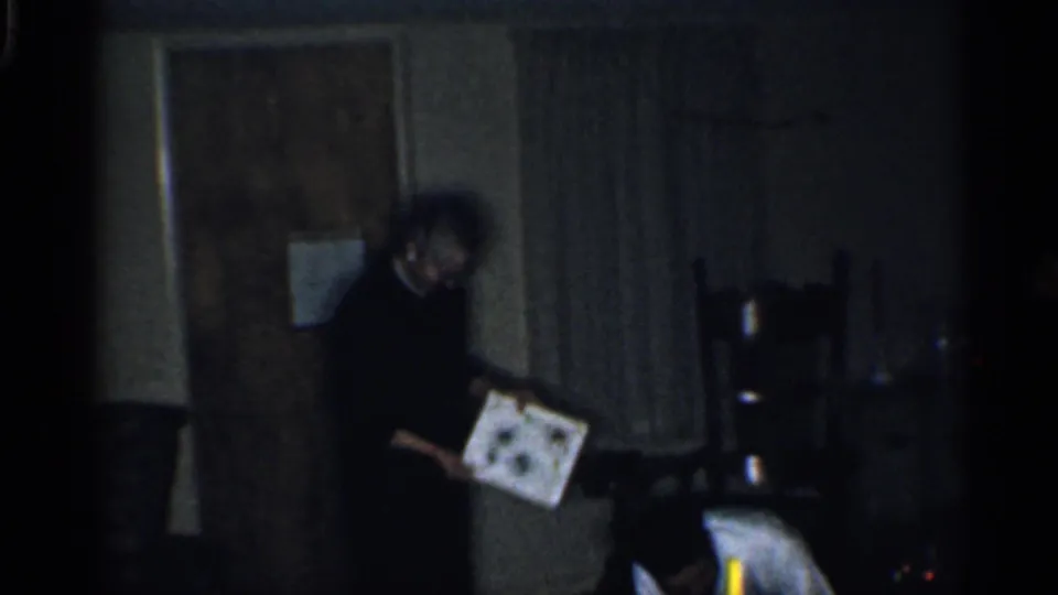 A person in a dark outfit stands near a doorway or curtain area, holding a picture in the dimly lit 1959 home interior.