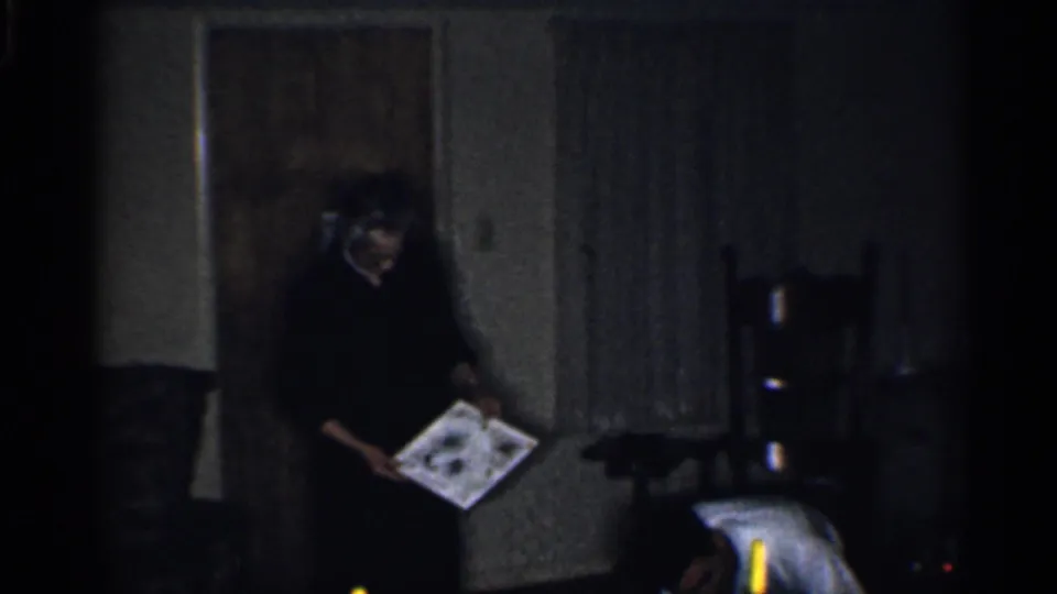 The person holding a picture is seen beside the doorway or curtain, with a chair and small candlelike light visible in the dark living room.