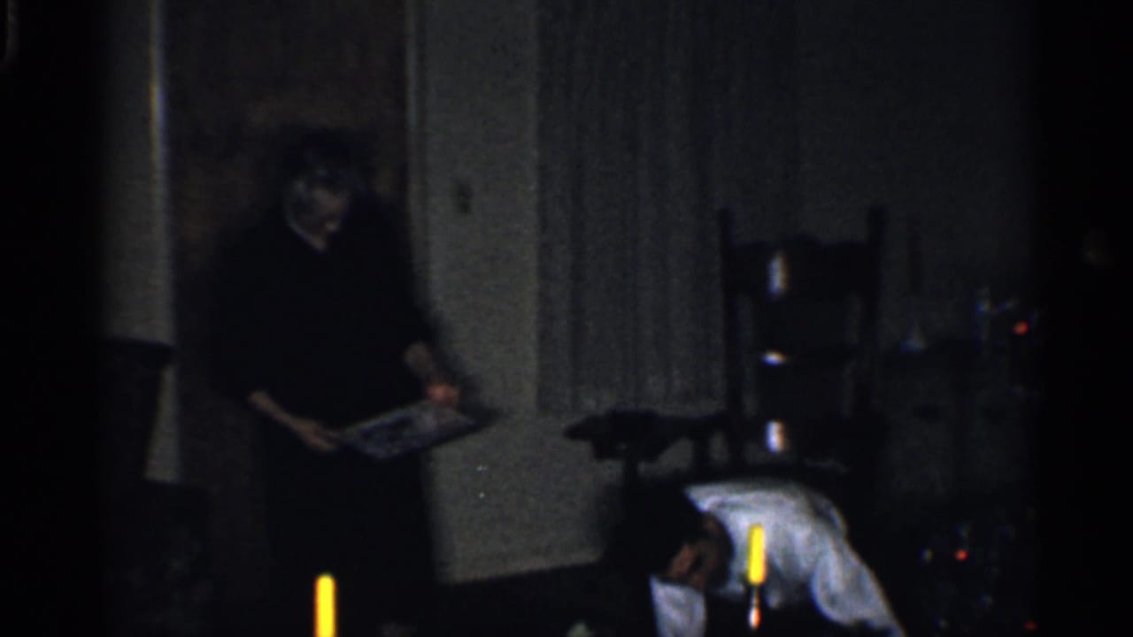 1959 California home-movie frame showing adults in a dark living room during Christmas gift preparation.