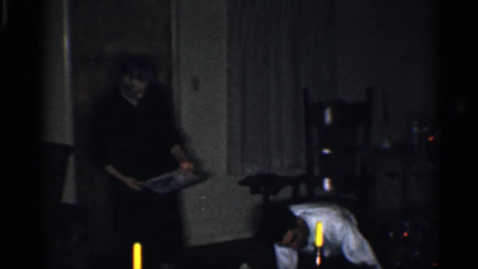 1959 California home-movie frame showing adults in a dark living room during Christmas gift preparation.