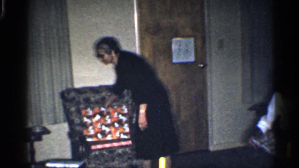 Home-movie frame of an adult beside a patterned armchair filled with Christmas gifts in a dim California living room.