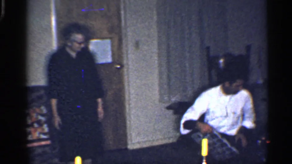 California 1959 home-movie frame with adults near wrapped presents in a dim living room holiday scene.