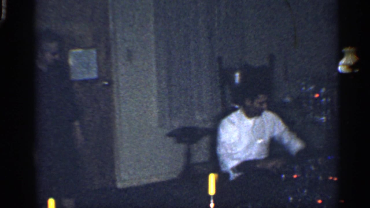 Adult in a white shirt seated in a dim room near a candle and doorway.