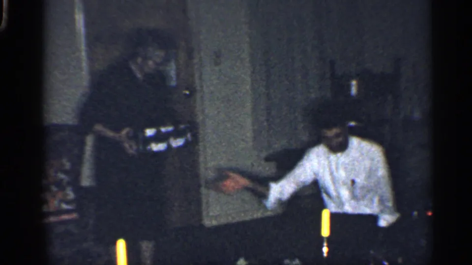 1959 California 8mm frame showing a woman in dark clothing offering a wrapped Christmas present to a seated adult in a dim room.