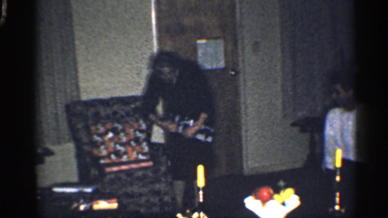Woman near a patterned chair opens a gift while a boy stands by the wall.