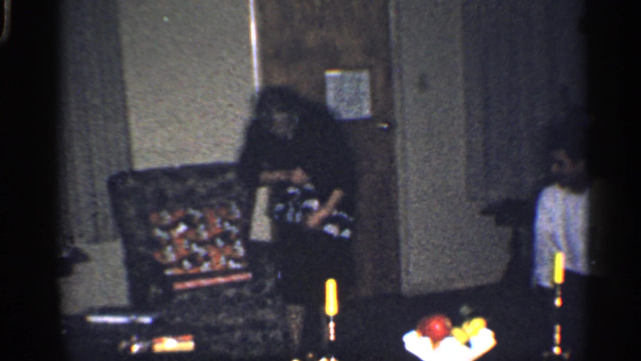 Woman continues unwrapping a gift near an armchair as a boy watches.