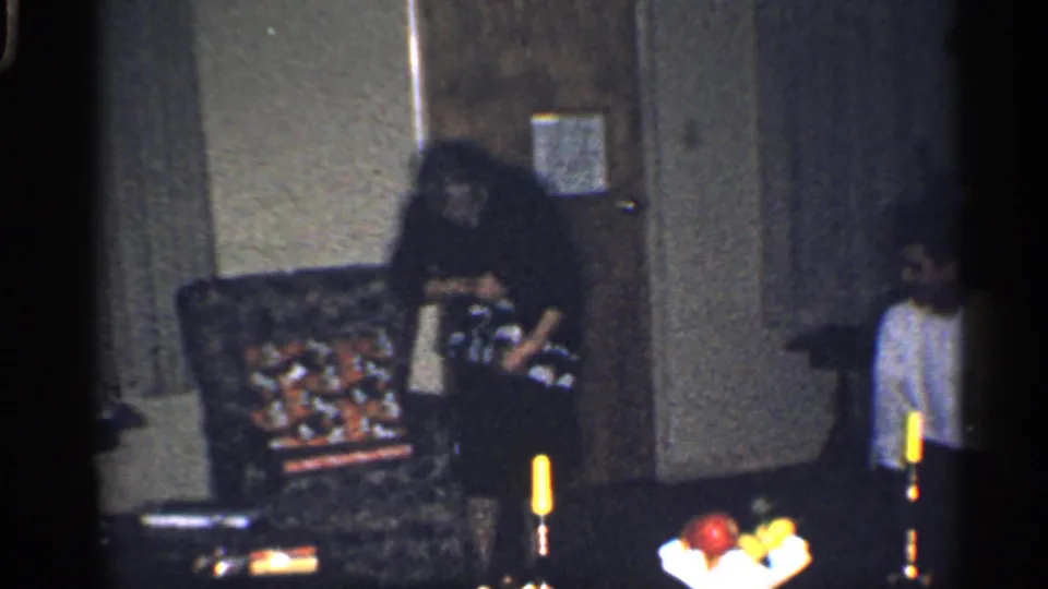 The woman works with the gift beside the armchair in the dim home interior, with the boy still standing near the wall.
